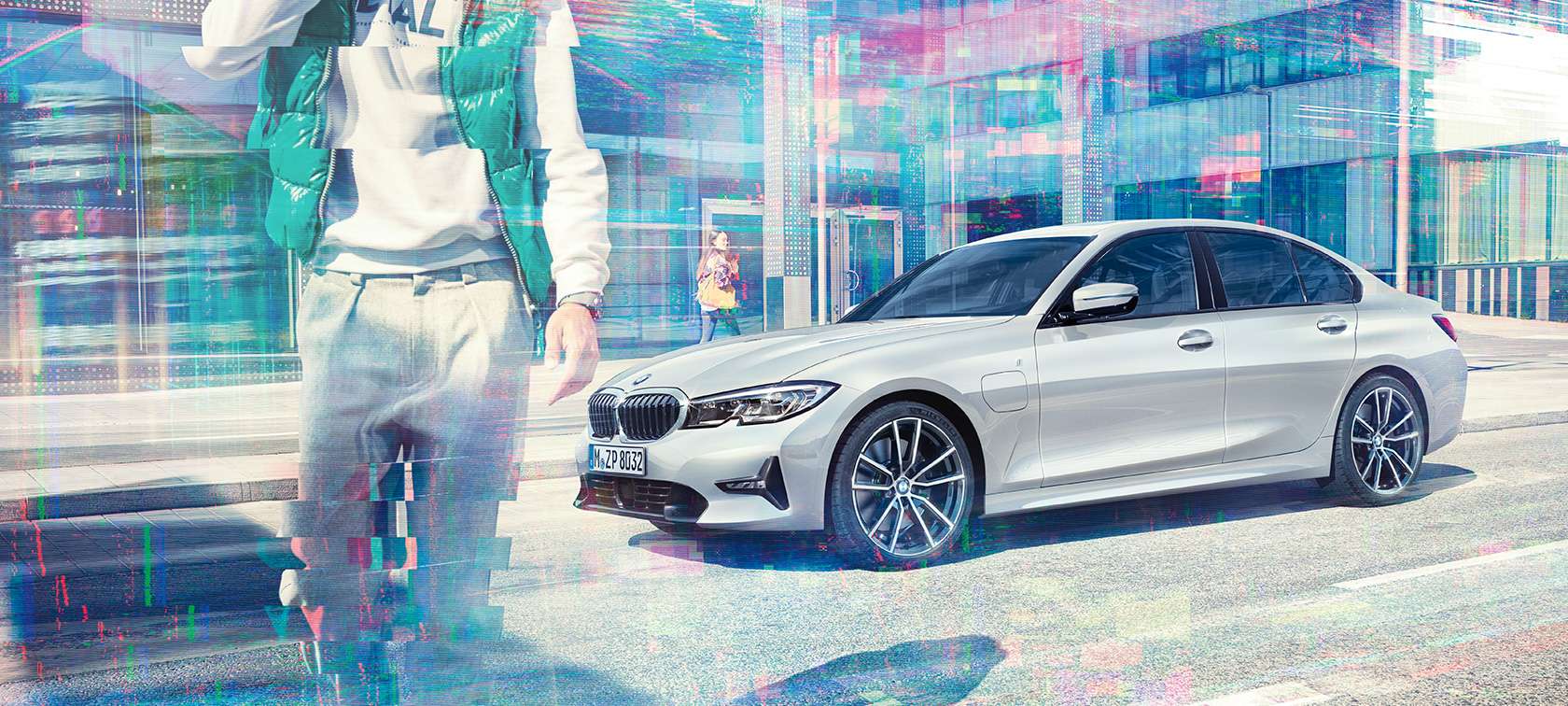 BMW Serije 3 Sedan, bočni snimak BMW 330e. Tročetvrtinski bočni snimak modela BMW 330e iPerformance Sedan koji stoji ispred urbane staklene zgrade sa potnicima koji hodaju.