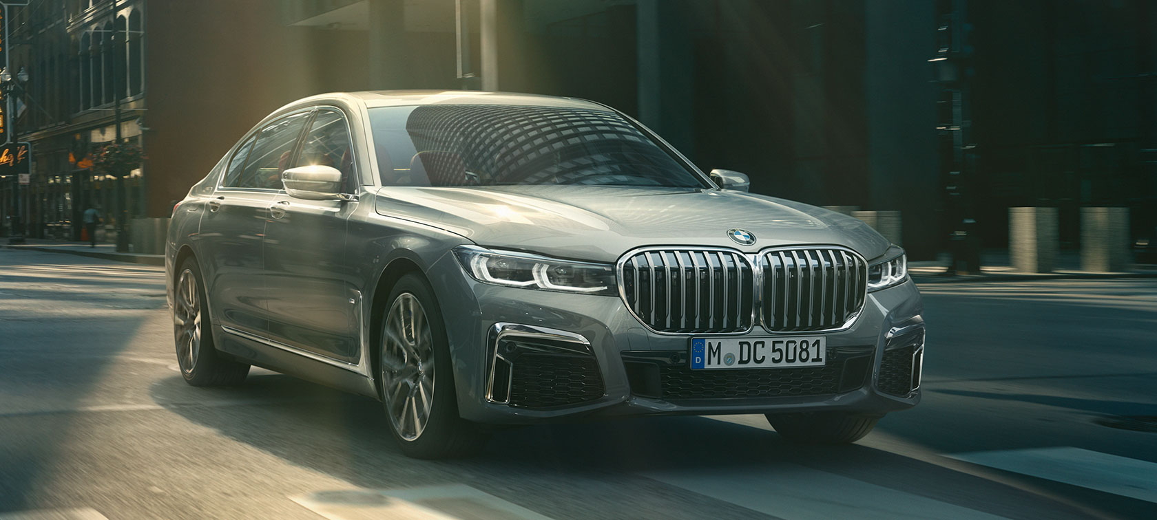 Sivi BMW Serije 7 Sedan u vožnji, tročetvrtinski pogled spreda Sivi BMW Serije 7 Sedan vozi kroz urbano okruženje u tročetvrtinskom pogledu spreda