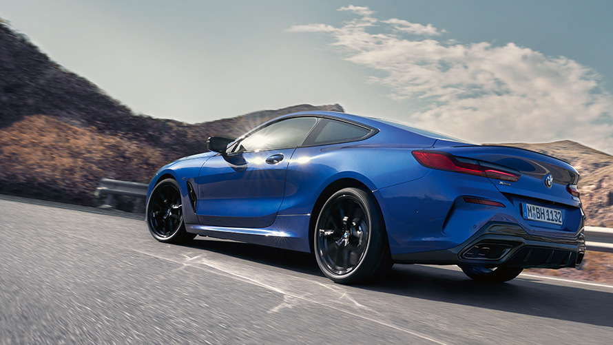 BMW Serije 8 Coupé G15 LCI Facelift tročetvrtinski zadnji prikaz vozila tokom vožnje BMW Serije 8 Coupé M Portimao plave metalik boje G15 LCI Facelift tročetvrtinski zadnji prikaz vozila prilikom vožnje sa brdima u pozadini