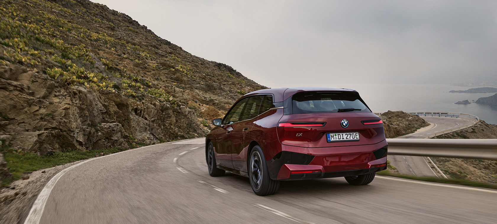 BMW iX i20 2021 električni SUV BMW iX xDrive50 vožnja pored obale BMW iX i20 2021 električni SUV BMW iX xDrive50 Aventurin crvene boje prikaz zadnjeg dela vozila na putu pored obale
