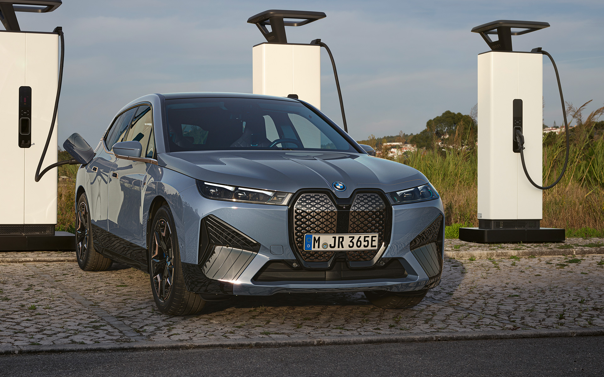 Prednji prikaz BMW-a iX M60 i20 2022 električni SUV BMW Individual Storm Bay metalik boje na stanici za punjenje velike snage BMW iX M60 i20 2022 električni SUV BMW Individual Storm Bay metalik prednji prikaz vozila na stanici za punjenje velike snage