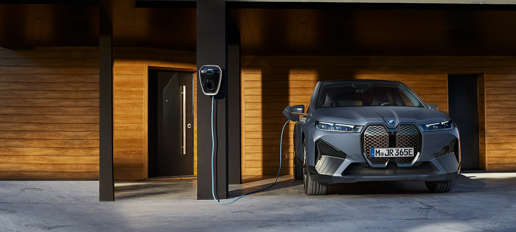 BMW iX M60 i20 2022 električni SUV električni automobil Oxid sive metalik boje prednji prikaz tokom punjenja ispred kuće preko BMW Wallbox-a 