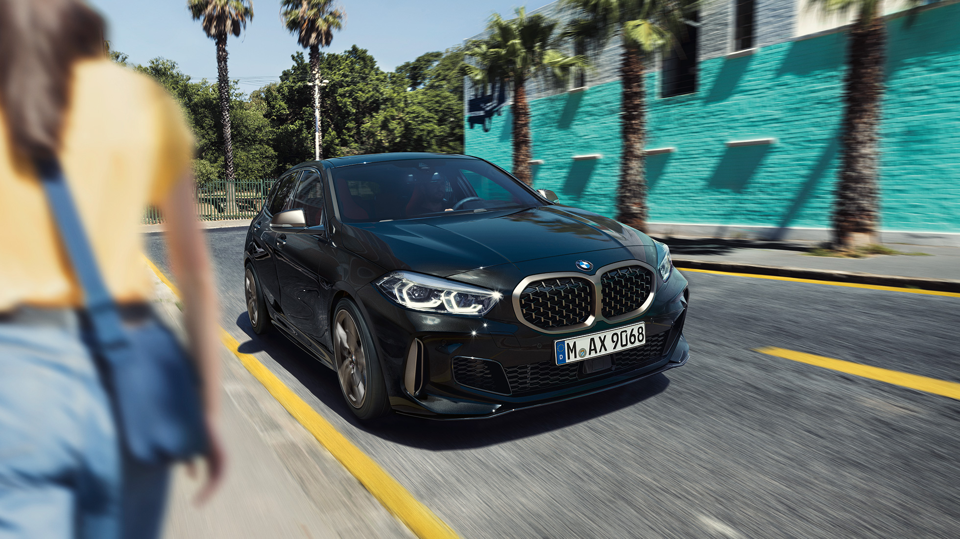 BMW M135i xDrive, three-quarter front view. BMW M135i xDrive u boji Black Sapphire metaliziranoj, tročetvrtinski pogled spreda u vožnji, sa ženom u prvom planu.