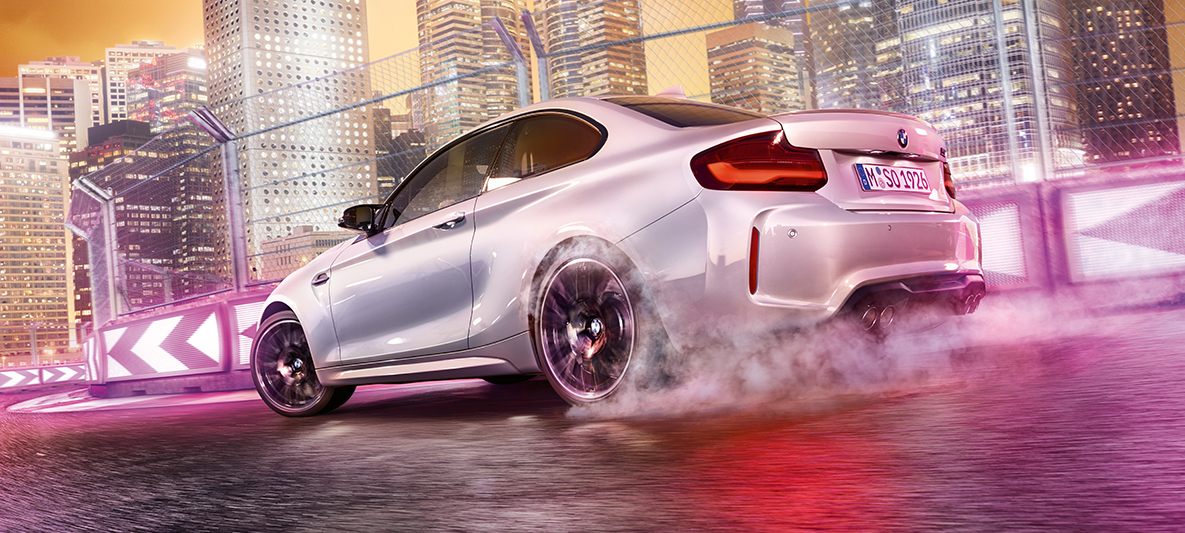 BMW M2 Competition ubrzava Beli BMW M2 Competition klizi po trkačkoj stazi ispred noćne gradske siluete.