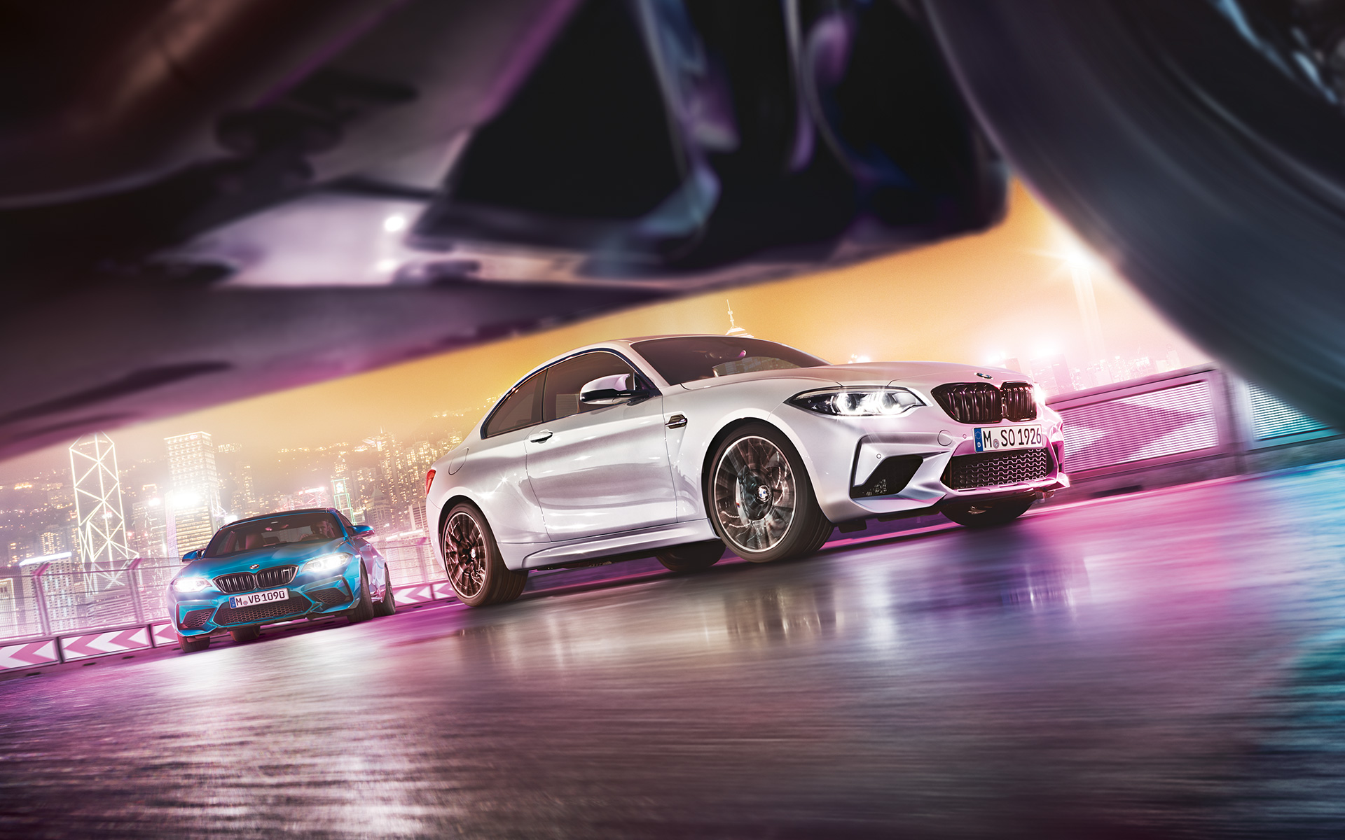 BMW M2 Competition na reflektivnom asfaltu. Dva modela BMW M2 Competition klize po trkačkoj stazi ispred noćnog gradskog pejzaža u nivou ulice.