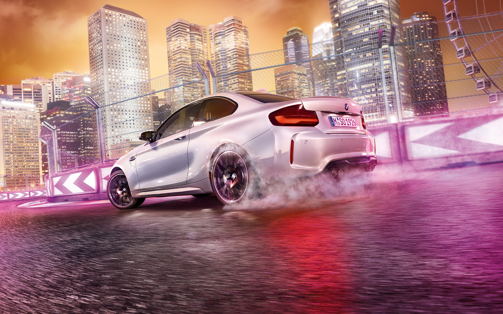 BMW M2 Competition ubrzava Beli BMW M2 Competition klizi po trkačkoj stazi ispred noćne gradske siluete.