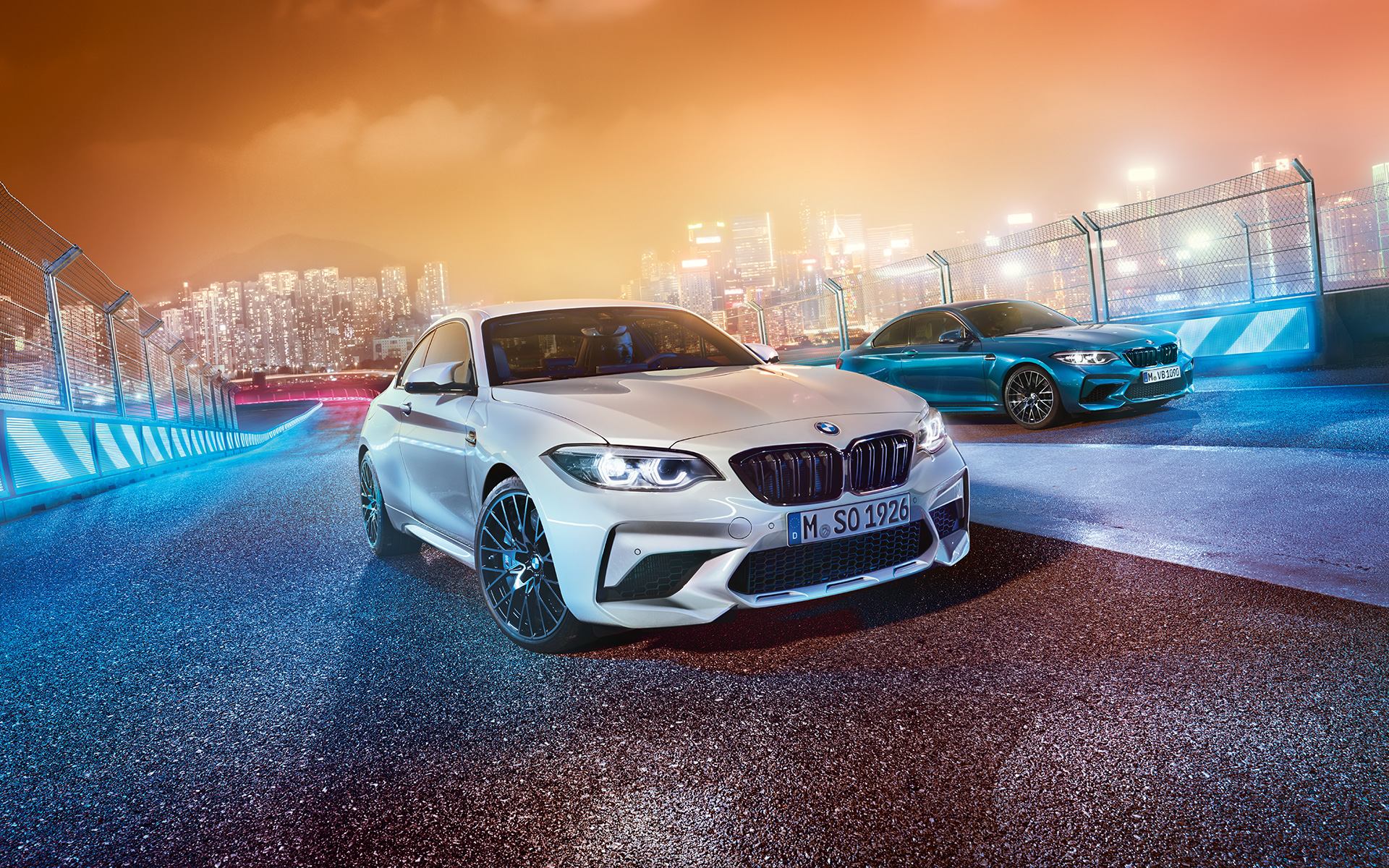 Dva modela BMW M2 Competition na trkačkoj stazi Beli i plavi BMW M2 Competition stoje na trkačkoj stazi ispred noćnog gradskog pejzaža.