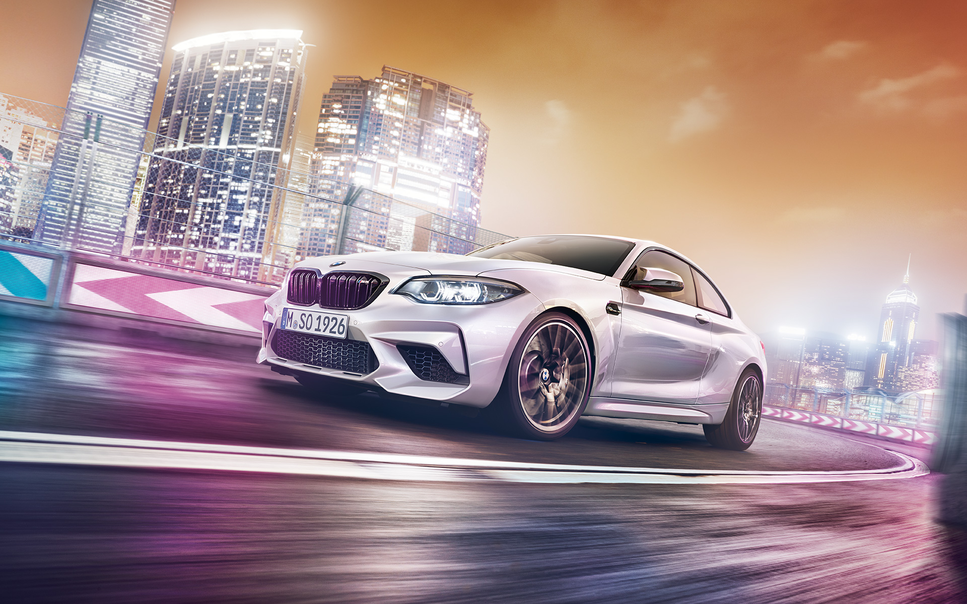 Beli BMW M2 Competition sa gradskom siluetom Beli BMW M2 Competition vozi po trkačkoj stazi ispred noćne gradske siluete.