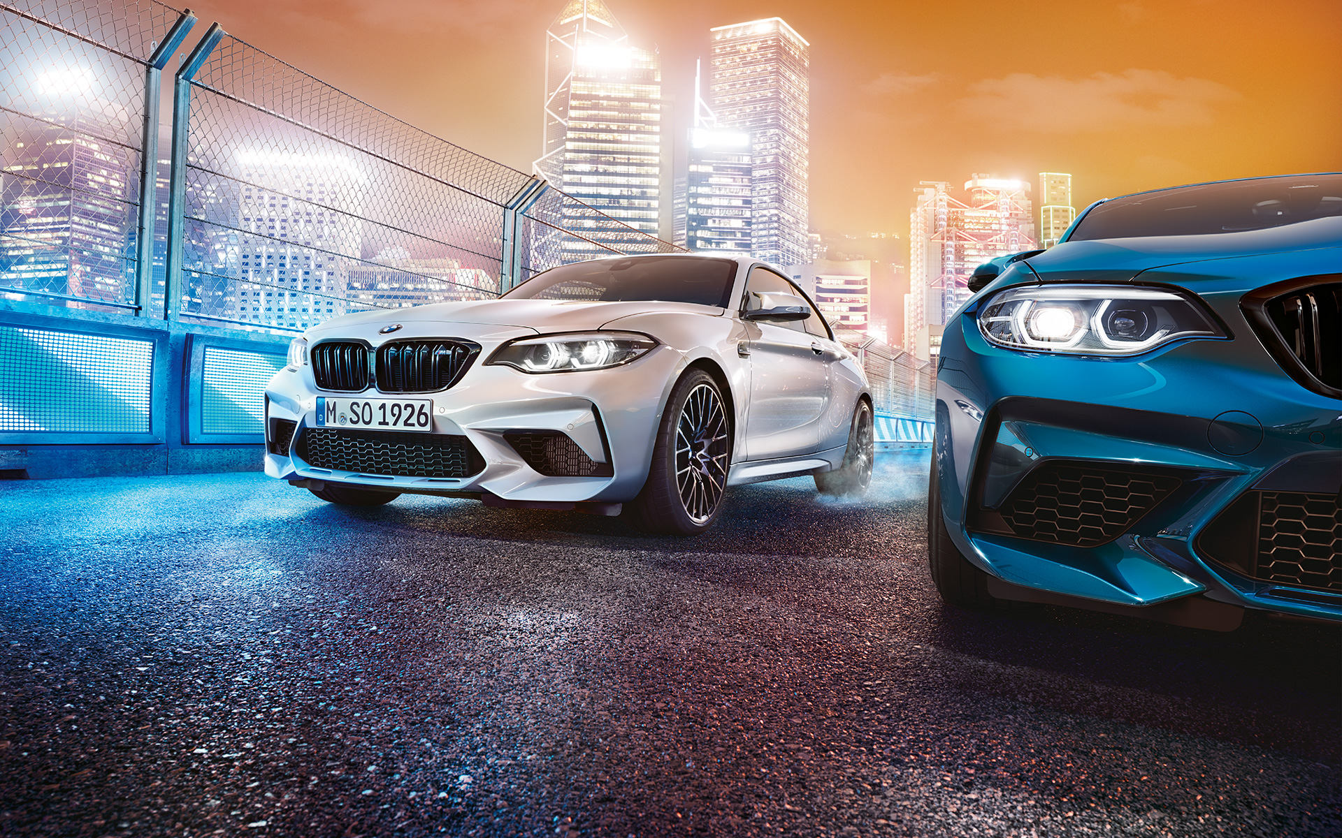 Beli i plavi BMW M2 Competition dok stoje Beli i plavi BMW M2 Competition spreda stoje ispred noćnog gradskog pejzaža.