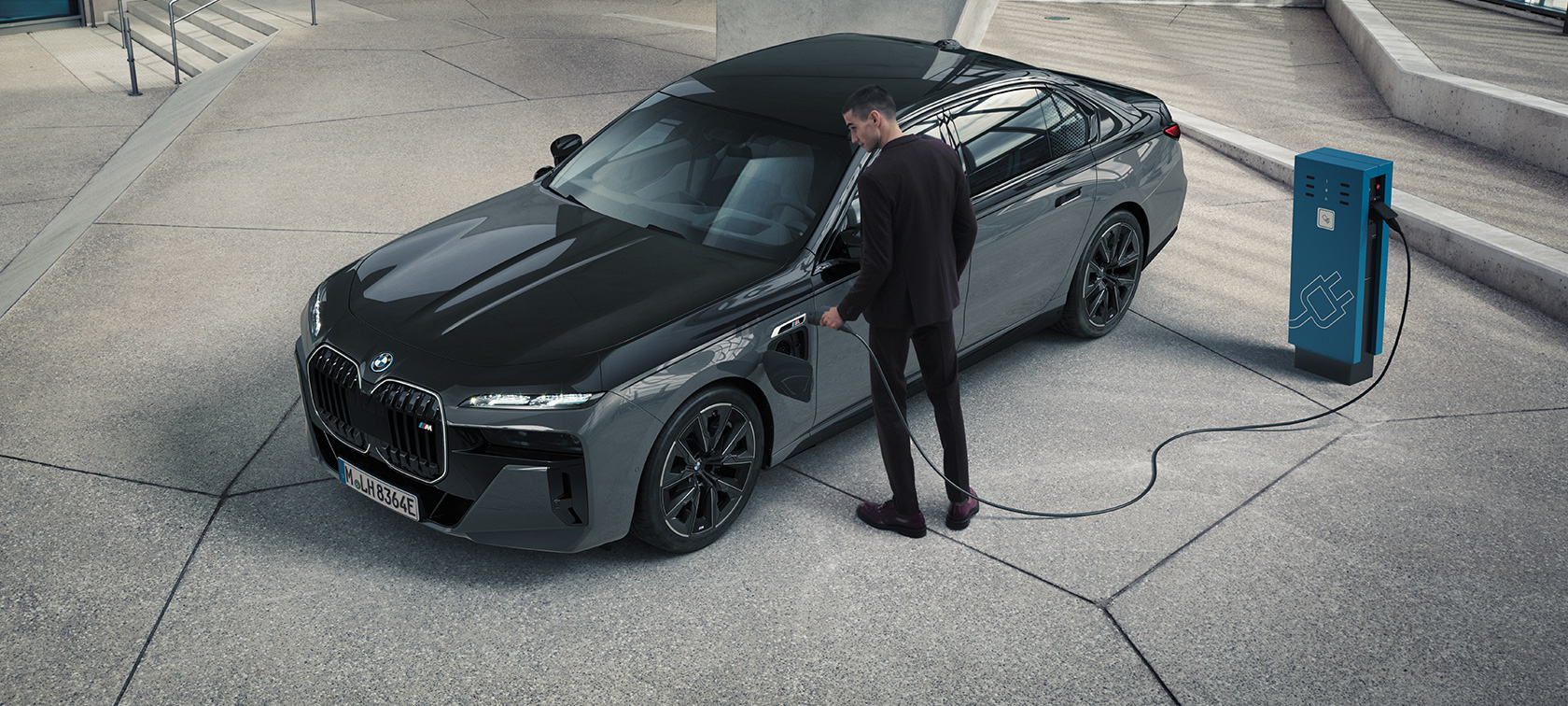 BMW M760e xDrive Sedan G70 M Performance paket BMW M760e xDrive Sedan G70 M Performance paket BMW Individual Black Sapphire metalik boja karoserije u dve nijanse BMW Individual Dravite siva metalik boja tročetvrtinski prednji prikaz čoveka s kablom za punjenje pored stanice za punjenje električnih vozila