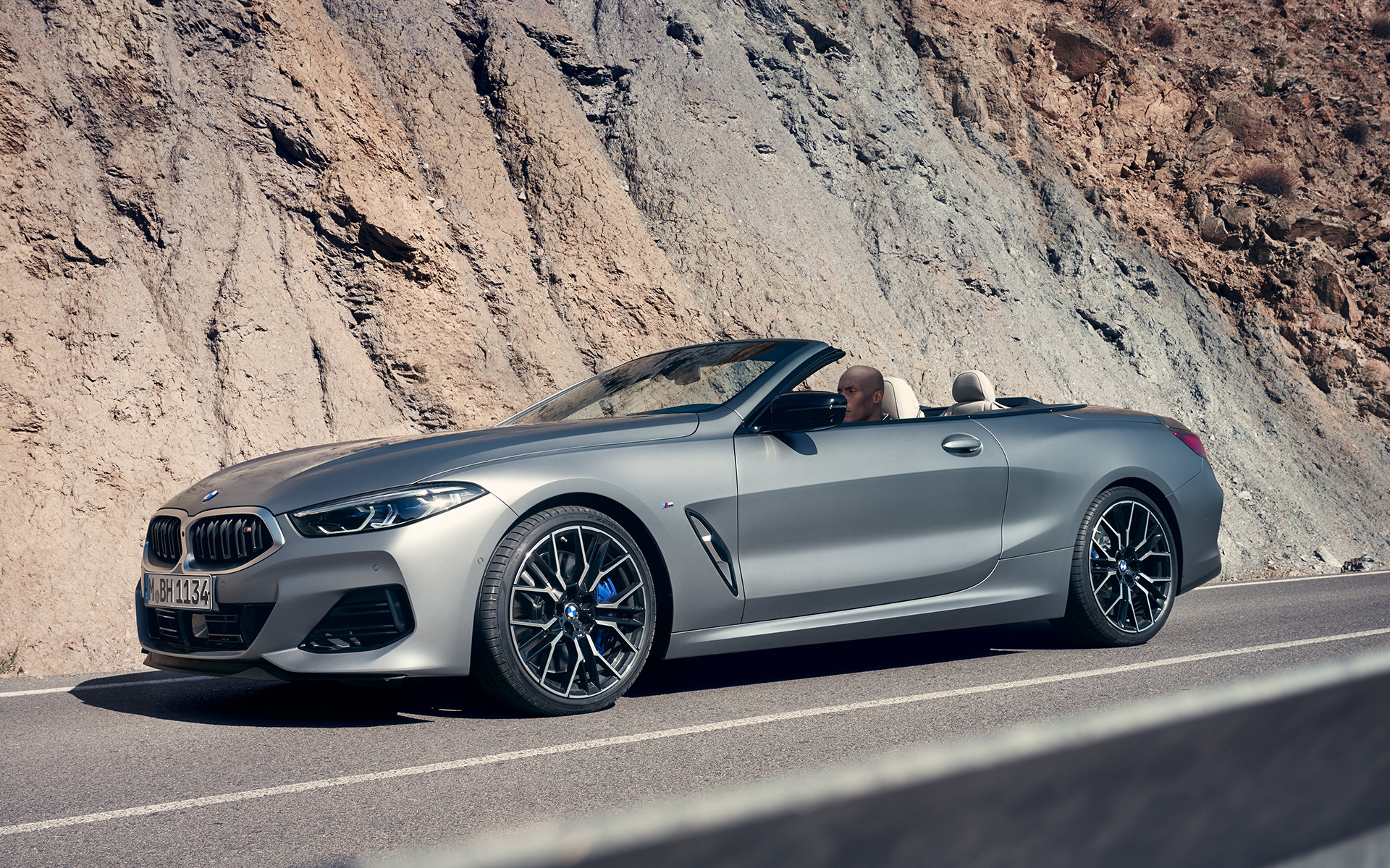 Tročetvrtinski prednji prikaz BMW M850i xDrive Kabriolet G14 LCI Facelift 2022 BMW M850i xDrive Kabriolet G14 LCI Facelift 2022 BMW Individual Frozen Pure siva metalik boja tročetvrtinski prednji prikaz vozila tokom vožnje s stenama u pozadini