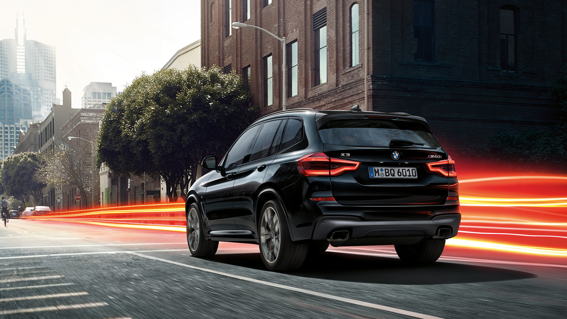 BMW X3 M40i, tročetvrtinski pogled otpozadi. BMW X3 M40i u boji Black Sapphire metaliziranoj, u vožnji, ejsterijer, tročetvrtinski pogled otpozadi ispred urbane pozadine sa svetlim prugama.
