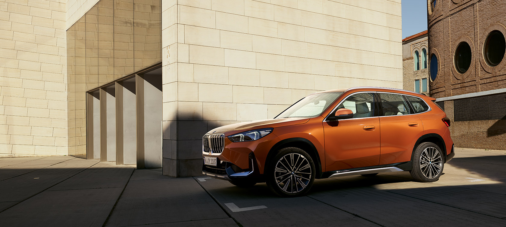 BMW U11 X1 xLine Utah narandžaste metalik boje tročetvrtinski prednji prikaz BMW U11 X1 xLine Utah narandžaste metalik boje tročetvtinski prednji prikaz nepomičnog vozila ispred zgrade