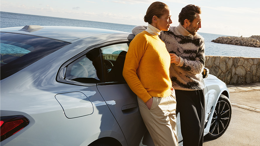 Trajanje usluge BMW Service Inclusive i ugovorni periodi za nove automobile BMW Service Inclusive muškarac i žena naslonjeni na modelu BMW i4 na obali