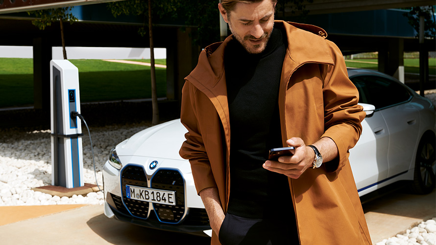 Trajanje usluge BMW Service Inclusive i ugovorni periodi za električna vozila BMW Service Inclusive čovek gleda u svoj pametni telefon ispred BMW-a i4 na stanici za punjenje